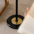 Close view of the round black marble base, brass disc and black stem of this Clérachic floor lamp, standing on a carpet.