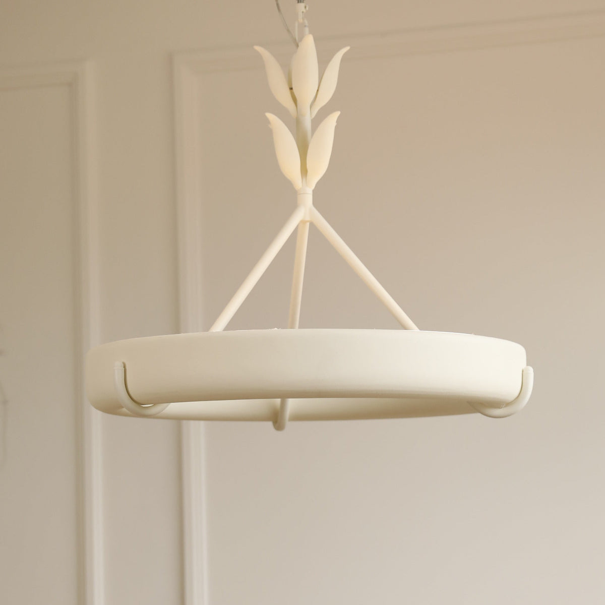 Front view of the white plaster ring held by three rods and a leaf-bud stem, against a warm beige wall, on a Clérachic chandelier.