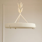 Side-on view of the matt white ring and its small curved hooks, hanging in front of a pale beige panelled wall; a Clérachic chandelier.