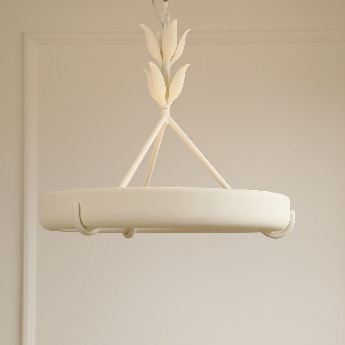 Side-on view of the matt white ring and its small curved hooks, hanging in front of a pale beige panelled wall; a Clérachic chandelier.