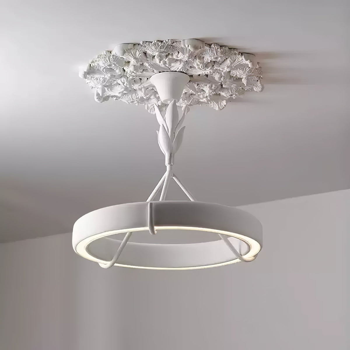 Lit white plaster ring below a ruffled plaster ceiling rose, with a soft glow along its inner edge; a Clérachic chandelier against a grey wall.