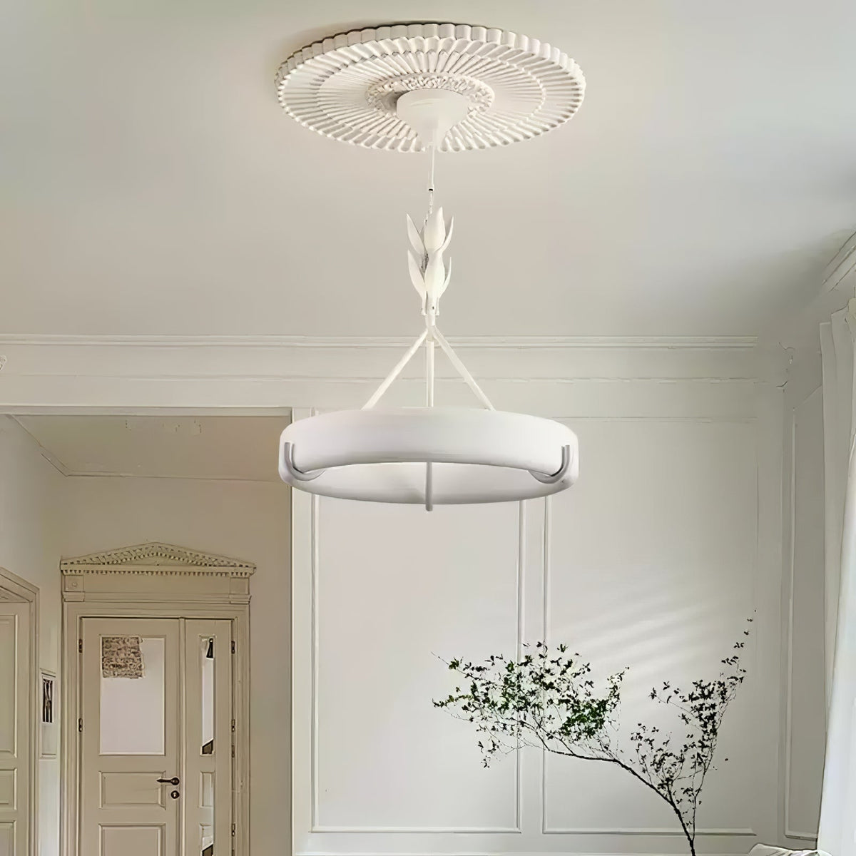 A round ceiling medallion with fine radial ridges sits above this Clérachic chandelier, in a room with a panelled wall, a door and a leafy branch.