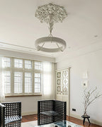From a distance, a Clérachic chandelier hangs under a carved plaster rose in a bright room with louvred shutters and black lattice armchairs.