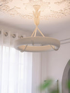 Upright photograph of the lit white ring and its leaf-bud stem below a plaster rose, in front of sheer curtains; a Clérachic chandelier.