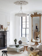 Tall view of a white living room with a window and gilded mirror, with this Clérachic chandelier hanging below a carved plaster rose.