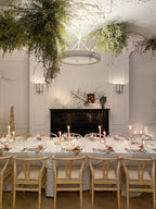 Hanging greenery crowds the ceiling around this Clérachic chandelier, above a long candlelit table with light wood chairs.