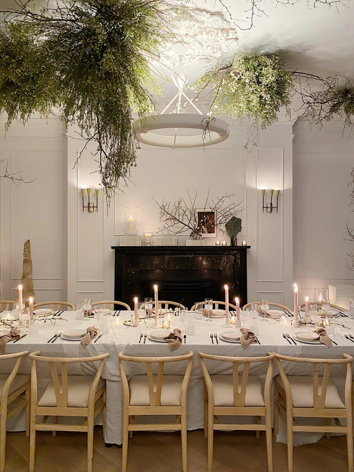 Hanging greenery crowds the ceiling around this Clérachic chandelier, above a long candlelit table with light wood chairs.