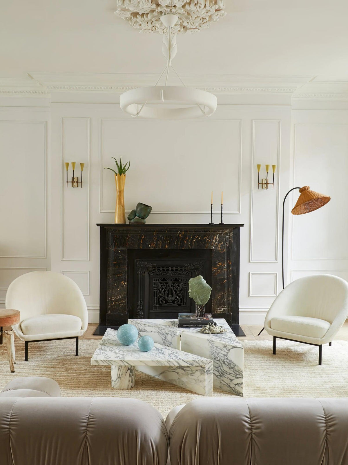 A Clérachic chandelier hangs in a symmetrical white room with two cream armchairs, a marble coffee table and brass wall lights either side of a fireplace.