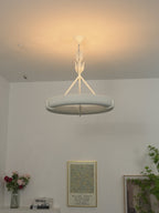 Warm ceiling glow around the ring and its leaf stem, with flowers and framed prints on a shelf below; a Clérachic chandelier.