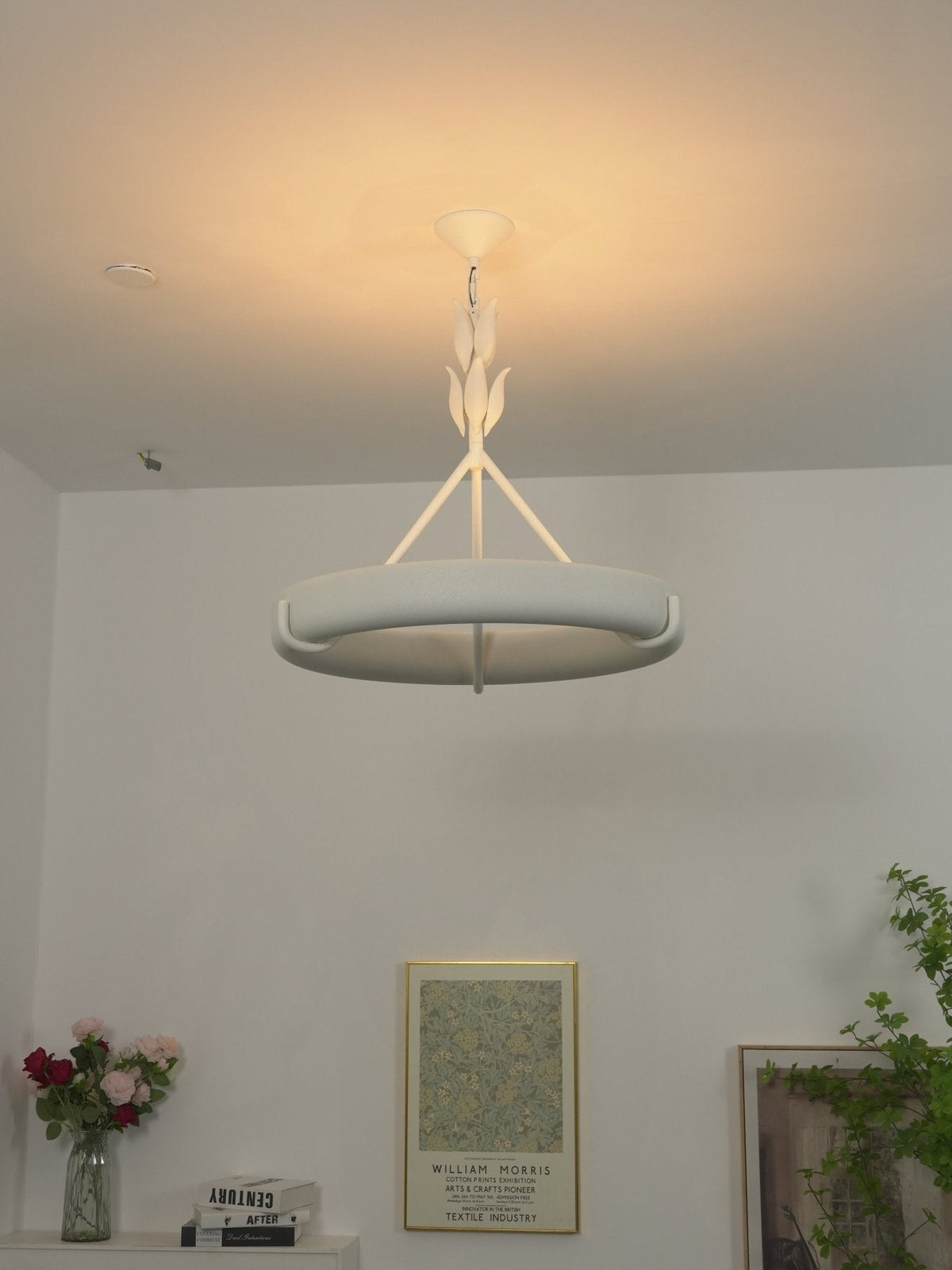 Warm ceiling glow around the ring and its leaf stem, with flowers and framed prints on a shelf below; a Clérachic chandelier.