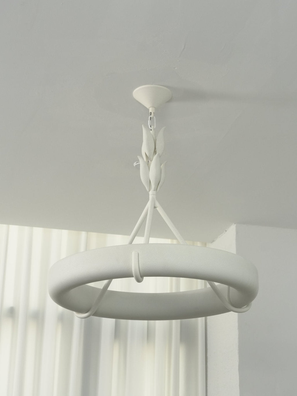 Cool daylight on the white ring, its leaf stem and ceiling cap, with curtains behind; a Clérachic chandelier.