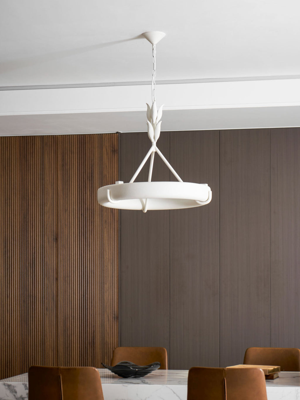 A Clérachic chandelier hangs in front of a ribbed wood wall and a marble table, with the chair backs just visible below.