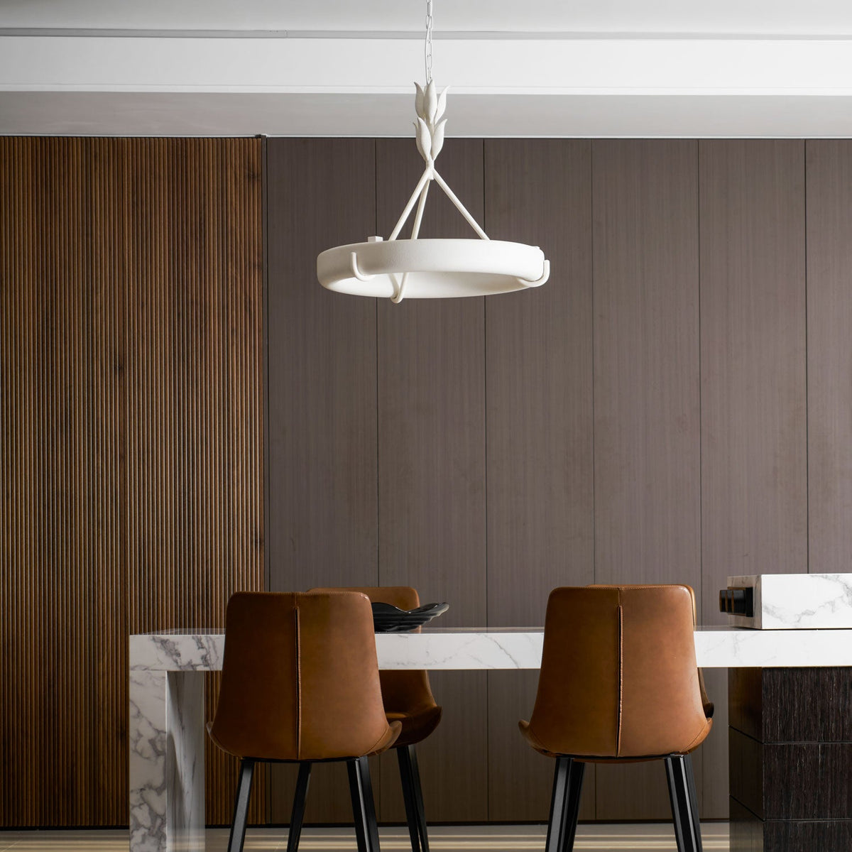 Slim white ring above a marble island and two tan leather chairs, with a ribbed wooden wall behind; a Clérachic chandelier.