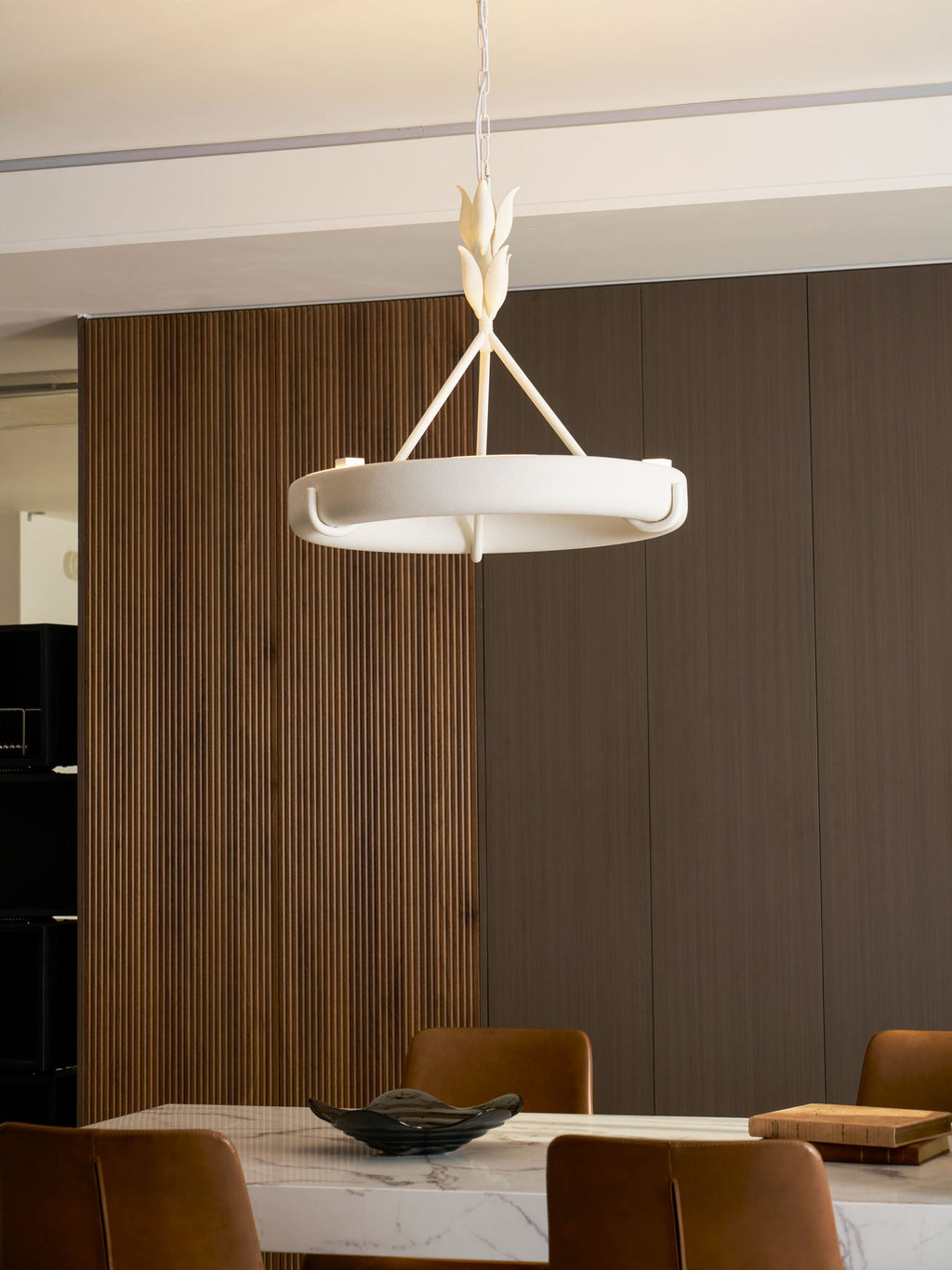 A Clérachic chandelier hangs beside a black appliance and a ribbed wooden panel, above a marble worktop with a wooden board.