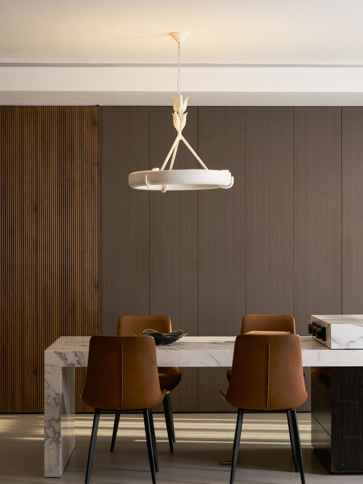 Pulled-back shot of the white ring above a marble dining table and two tan leather chairs with black legs; a Clérachic chandelier.