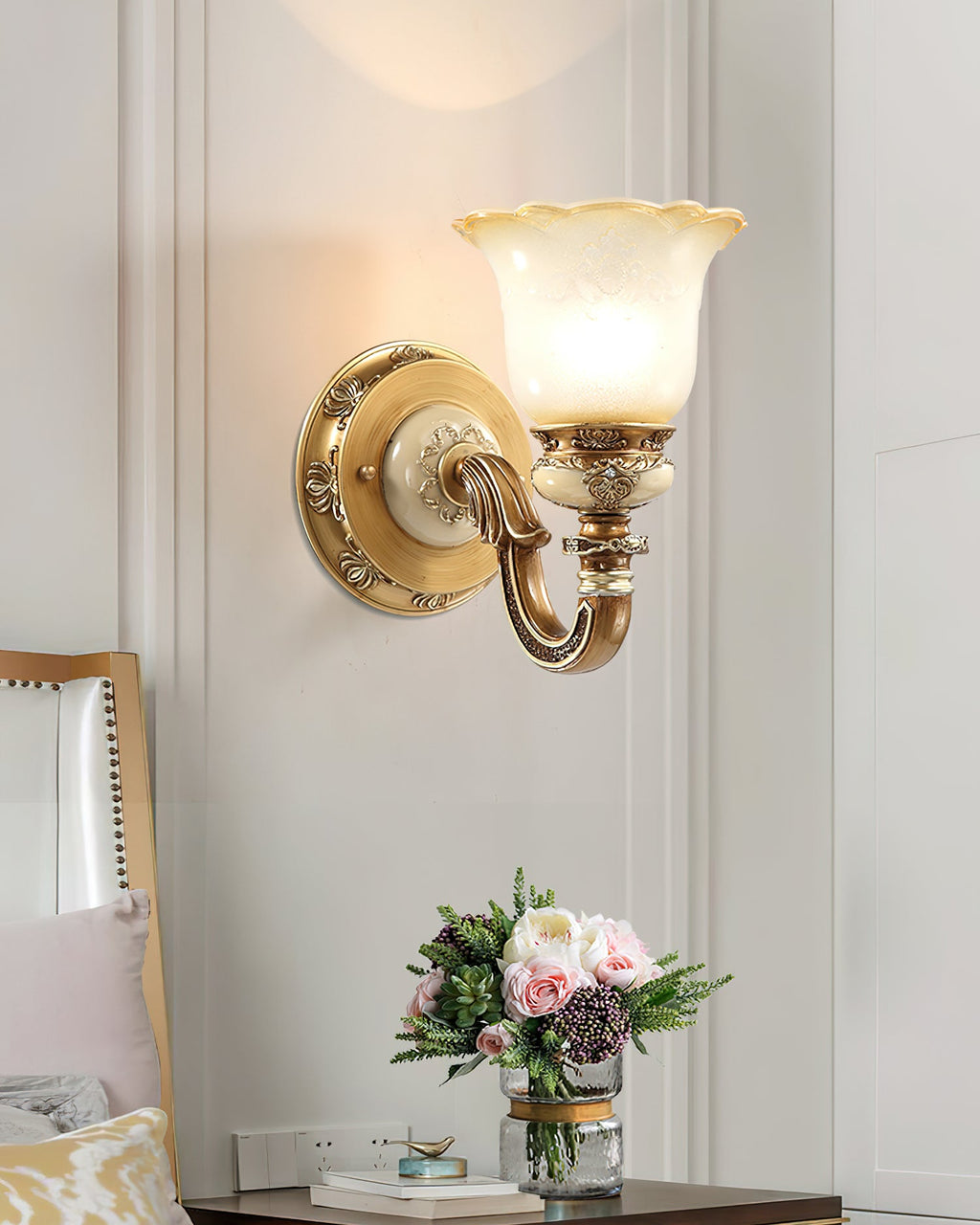 Single-head Clérachic wall light glowing on a white panelled wall above a small bedside table and a posy of pink flowers.