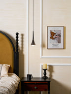 A small Clérachic pendant glows warm beside a bed with a mustard headboard, above a dark bedside table with a mug.