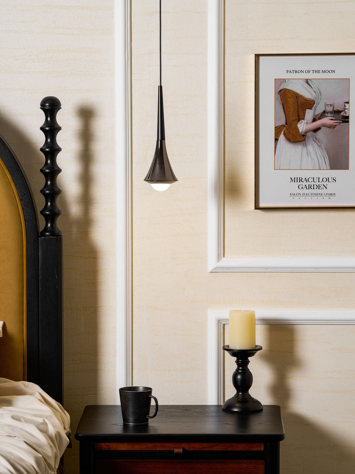 A slender black Clérachic pendant with an opal bulb hangs beside a bedpost, above a bedside table with a candle.