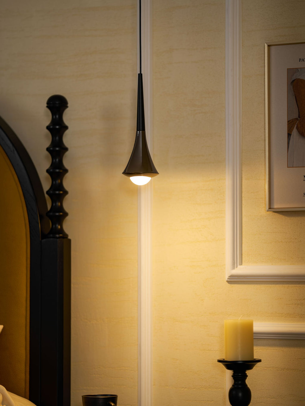 A black Clérachic pendant gives a warm light to a cream panelled wall, with a black bedpost and a candle below.