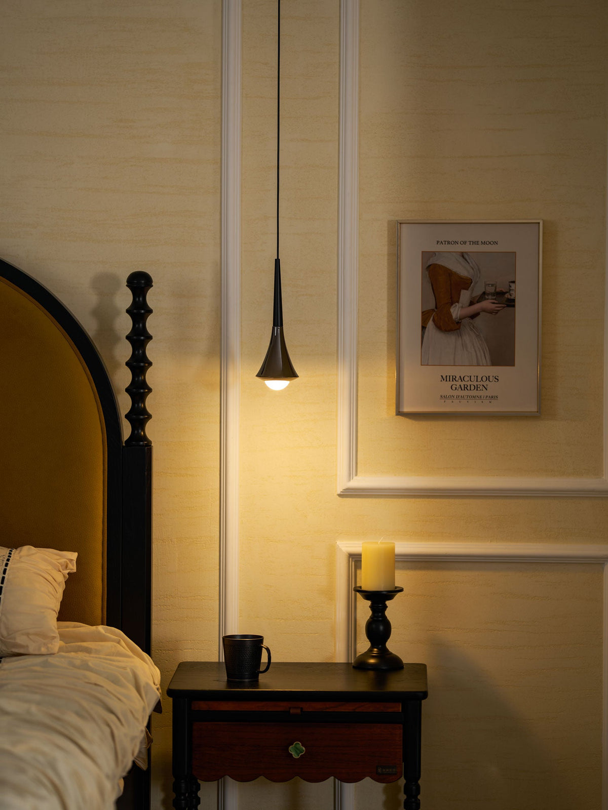 Warm evening light from a slim black Clérachic pendant beside a bed, with a framed poster and a candle on the table.
