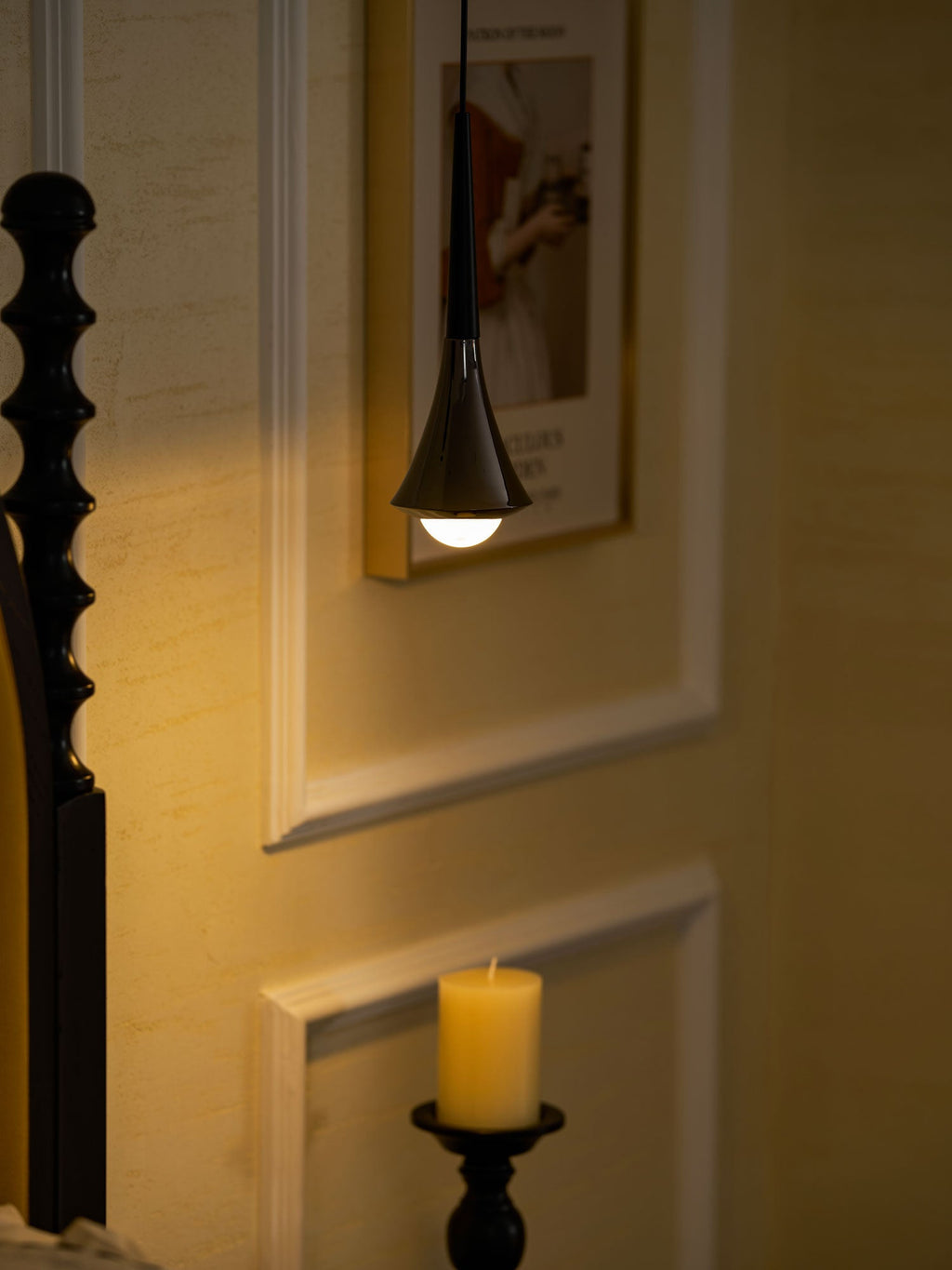 A black Clérachic pendant glows warm against a golden panelled wall, with a carved bedpost and a candle nearby.
