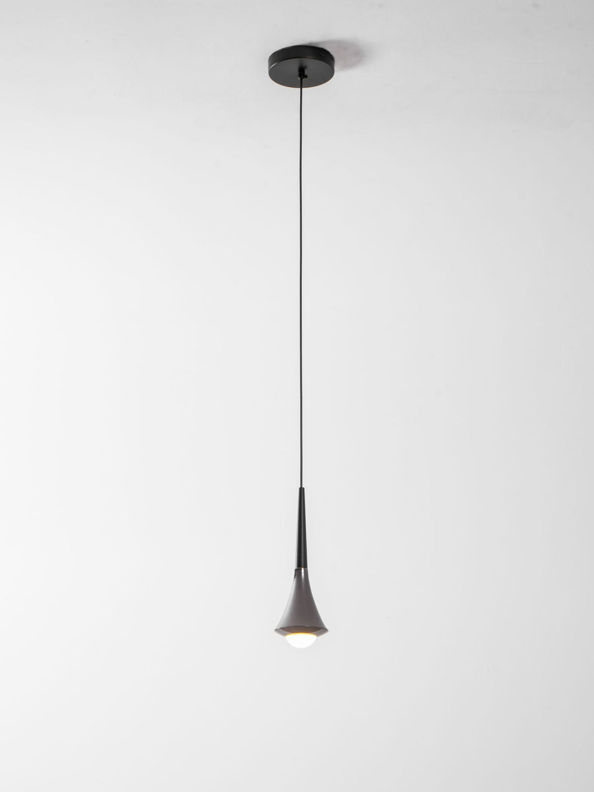 A Clérachic pendant on a long thin cord from a small black canopy, with a lit opal bulb at the tip, on a pale backdrop.