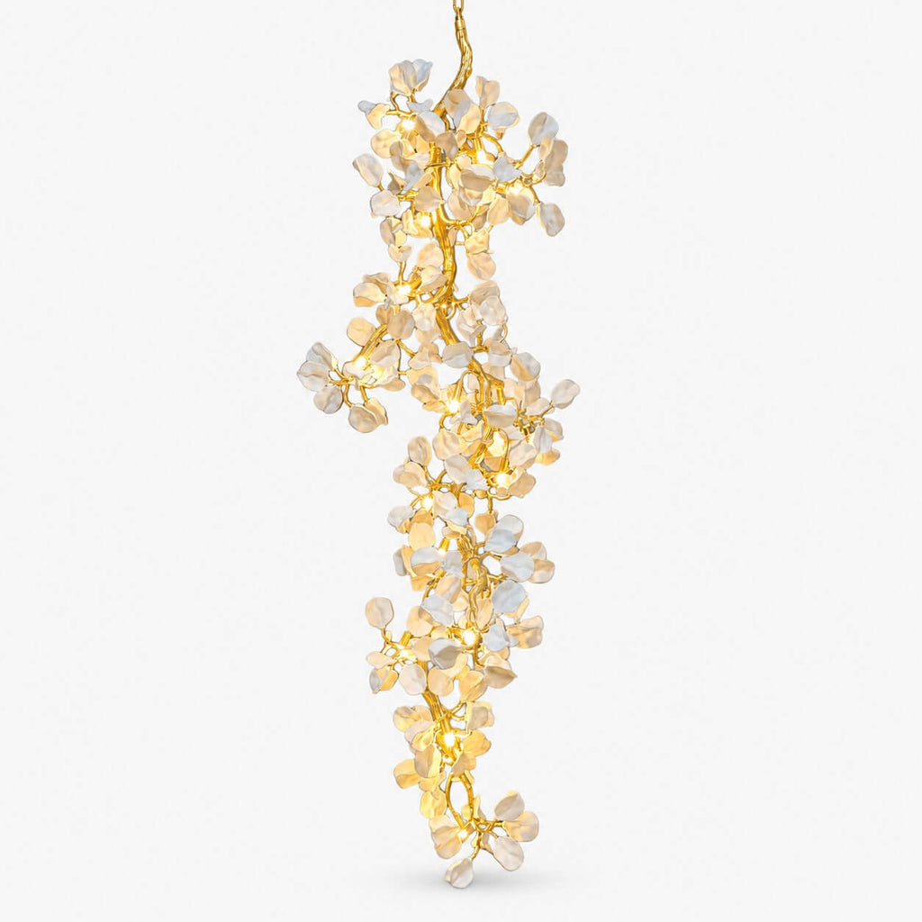 Cascading chandelier from Clérachic, a long column of pale ceramic petals on a gold vine frame, lit warmly on a plain white backdrop.