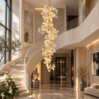 Long Clérachic ceramic petal chandelier hanging beside a sweeping white staircase with a glass balustrade in a marble-floored hall.