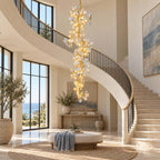 Clérachic gold and ceramic petal chandelier beside a curved staircase with an iron handrail, with a cream pouffe and a woven rug below.