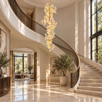 A Clérachic ceramic petal chandelier beside a sweeping stairwell, with a marble floor and large olive trees in pots.