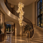 A Clérachic petal chandelier hung low beside a curved staircase and tall windows, in a warm-lit entrance hall with a marble floor.