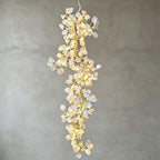 Lit Clérachic cascading chandelier of ceramic petals and gold-tone branches against a grey plaster wall.