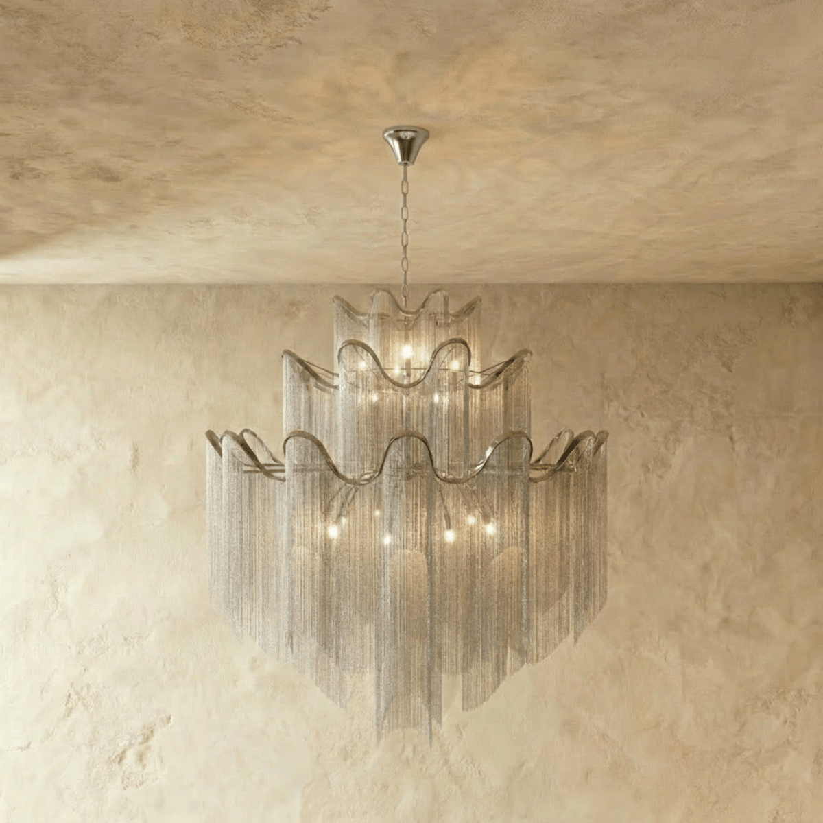 Clérachic silver three-tier chandelier with long chain fringe, shown lit and hung from a chain before a plaster wall.