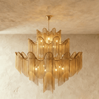 Clérachic larger gold three-tier chandelier with a wide spread of chain fringe, lit and hung against a plaster wall.