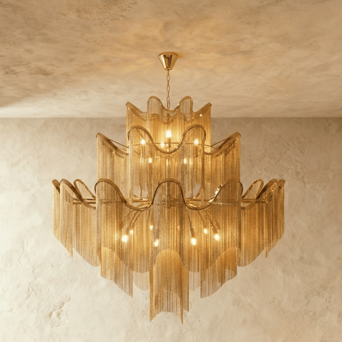 Clérachic larger gold three-tier chandelier with a wide spread of chain fringe, lit and hung against a plaster wall.