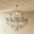 Clérachic larger silver three-tier chandelier with a wide spread of chain fringe, lit and hung against a plaster wall.