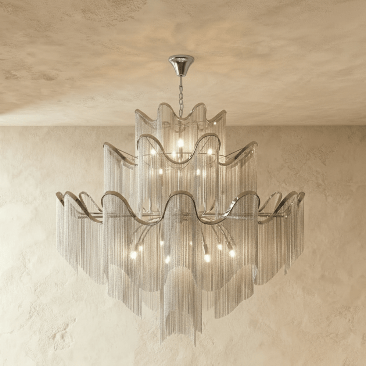 Clérachic larger silver three-tier chandelier with a wide spread of chain fringe, lit and hung against a plaster wall.