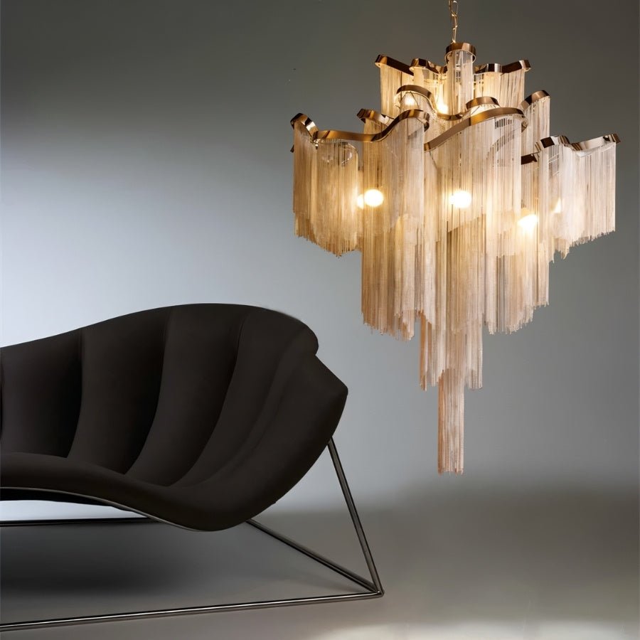 Clérachic chain-fringe chandelier glowing warm gold above a black sculptural lounge chair on a grey studio backdrop.