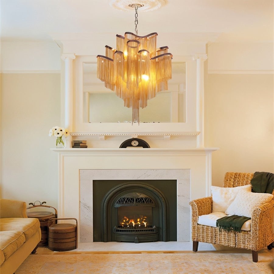 Clérachic gold chain-fringe chandelier hanging above a white fireplace and mantel, with a mirror and wicker armchair beside it.