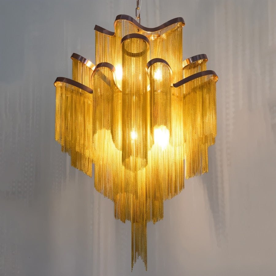 Clérachic gold chain-fringe chandelier close up, lit warmly, with its long chain tails and curved ribbon rims against a grey wall.