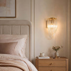 This lit Clérachic wall light hangs above an oak bedside table with a small vase, beside an upholstered headboard and pink bedding.