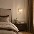 Over a dark wood nightstand with white roses, a Clérachic wall light with clear glass strands glows beside a panelled bedroom wall.