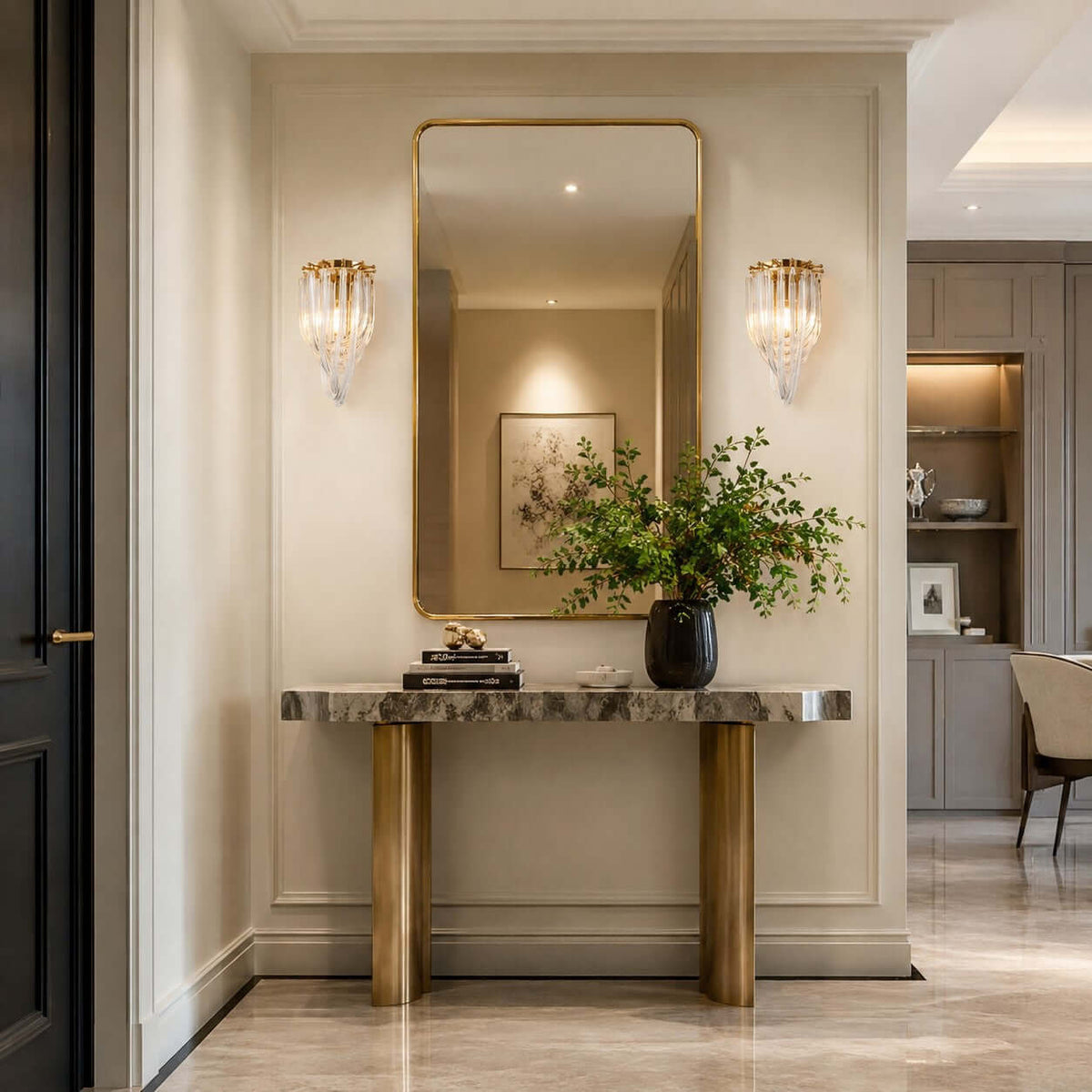 A pair of Clérachic wall lights flank a tall gold-framed mirror above a marble console with brass legs, in a cream hallway.