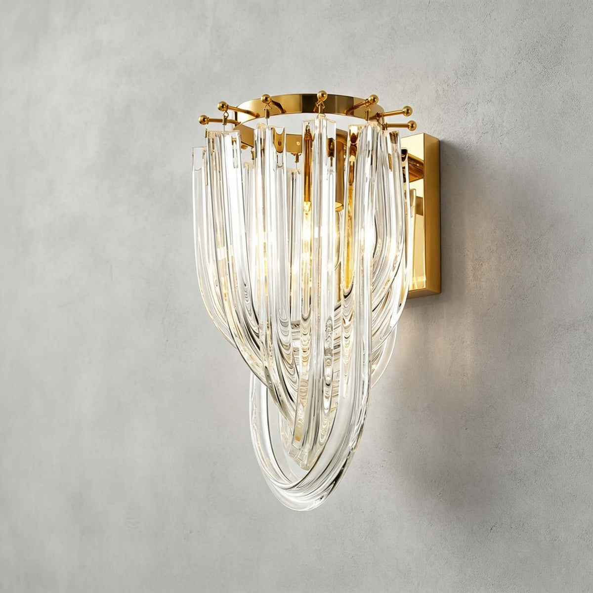 Close view of the looped clear glass strands and the gold backplate of this Clérachic wall light, on a grey plaster wall.