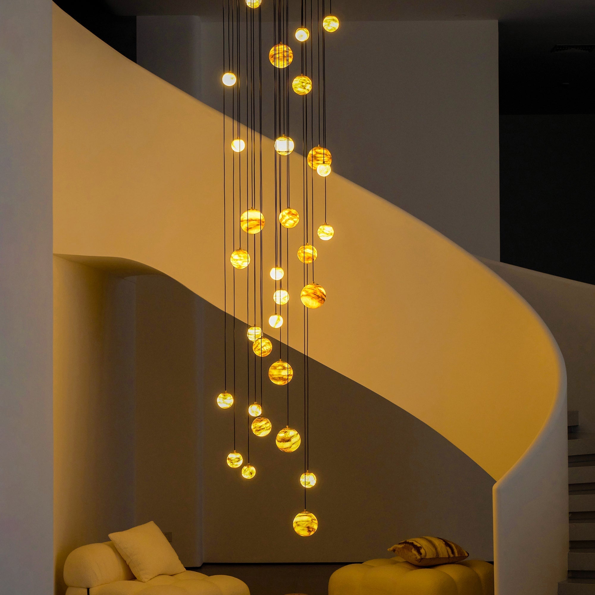 The Clérachic Cascading Globe Chandelier after dark, lit spheres washing the curved stair wall in amber against a darkened room.
