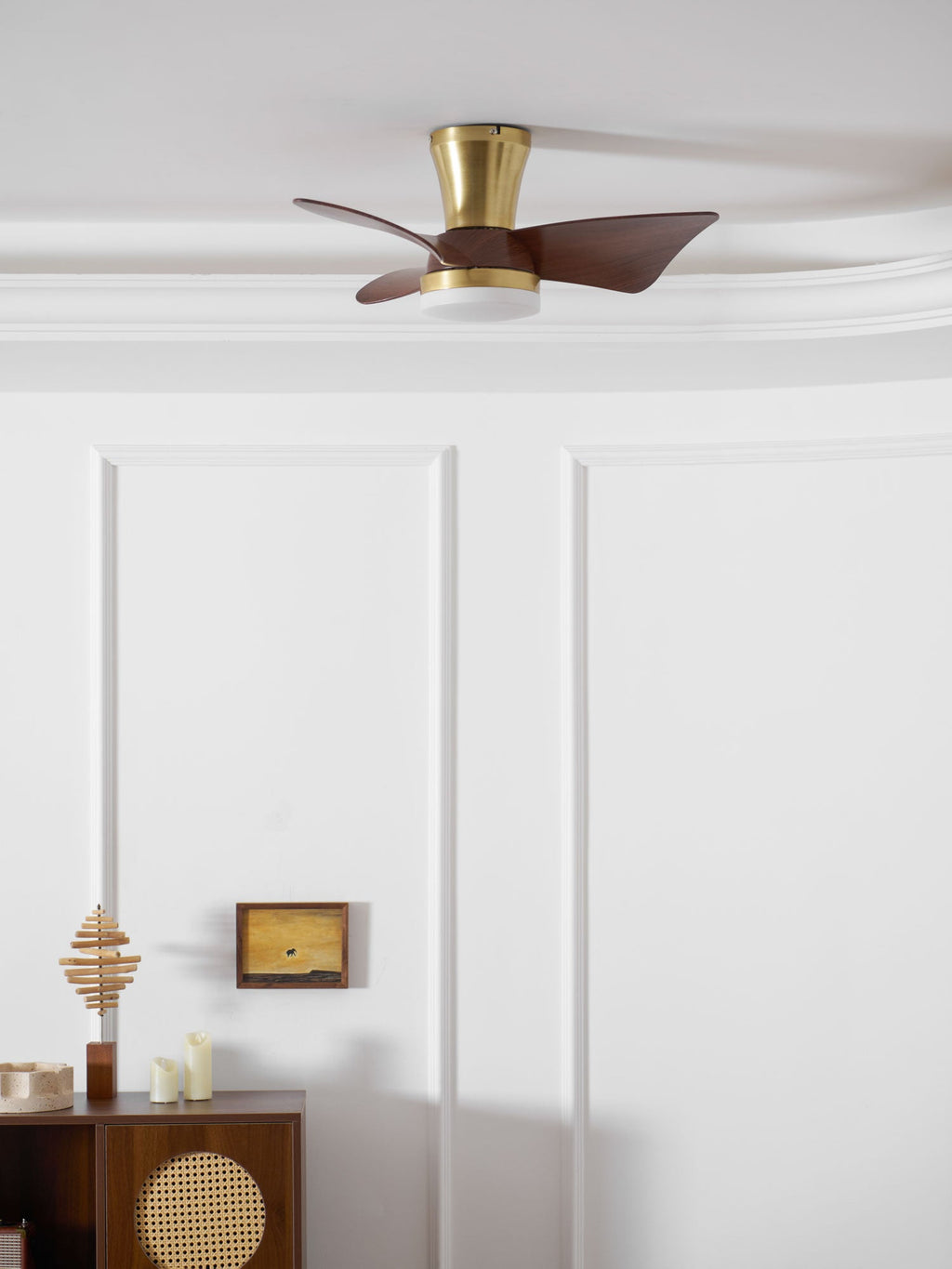 A Clérachic gold ceiling fan with three dark blades above a white panelled wall, with a framed picture and a cane-fronted cabinet below.