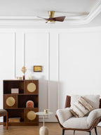 A wide room view with the Clérachic gold ceiling fan overhead, a wooden shelf unit, a small side table and a cream armchair below.