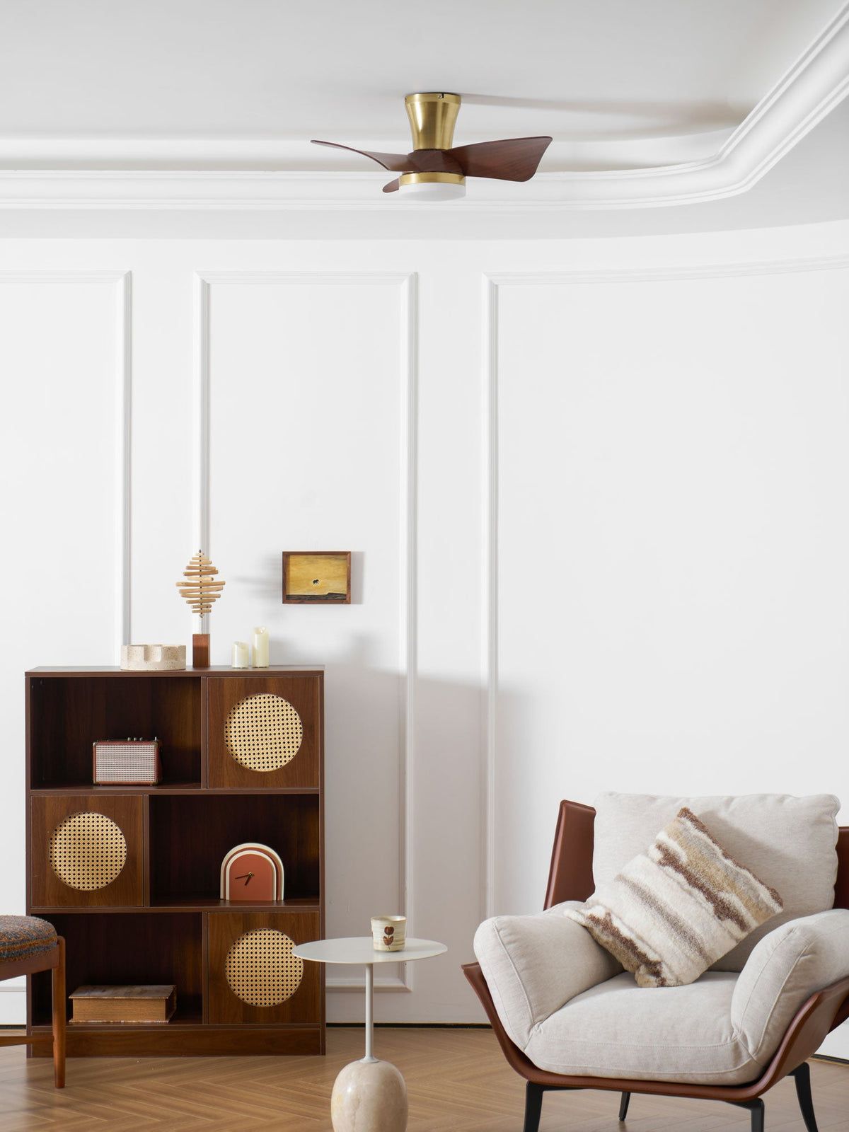 A wide room view with the Clérachic gold ceiling fan overhead, a wooden shelf unit, a small side table and a cream armchair below.