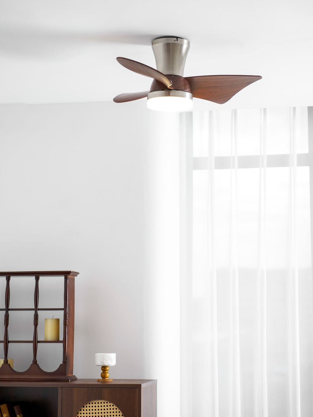 A Clérachic nickel and walnut ceiling fan with a lit light, in a bright room with sheer curtains and a dark wooden display cabinet.
