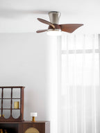 A Clérachic nickel and walnut ceiling fan with a lit light, in a bright room with sheer curtains and a dark wooden display cabinet.